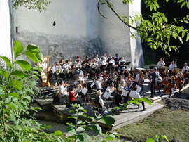 Das Orchester sitzt im Freien vor einem Steingebäude und probt